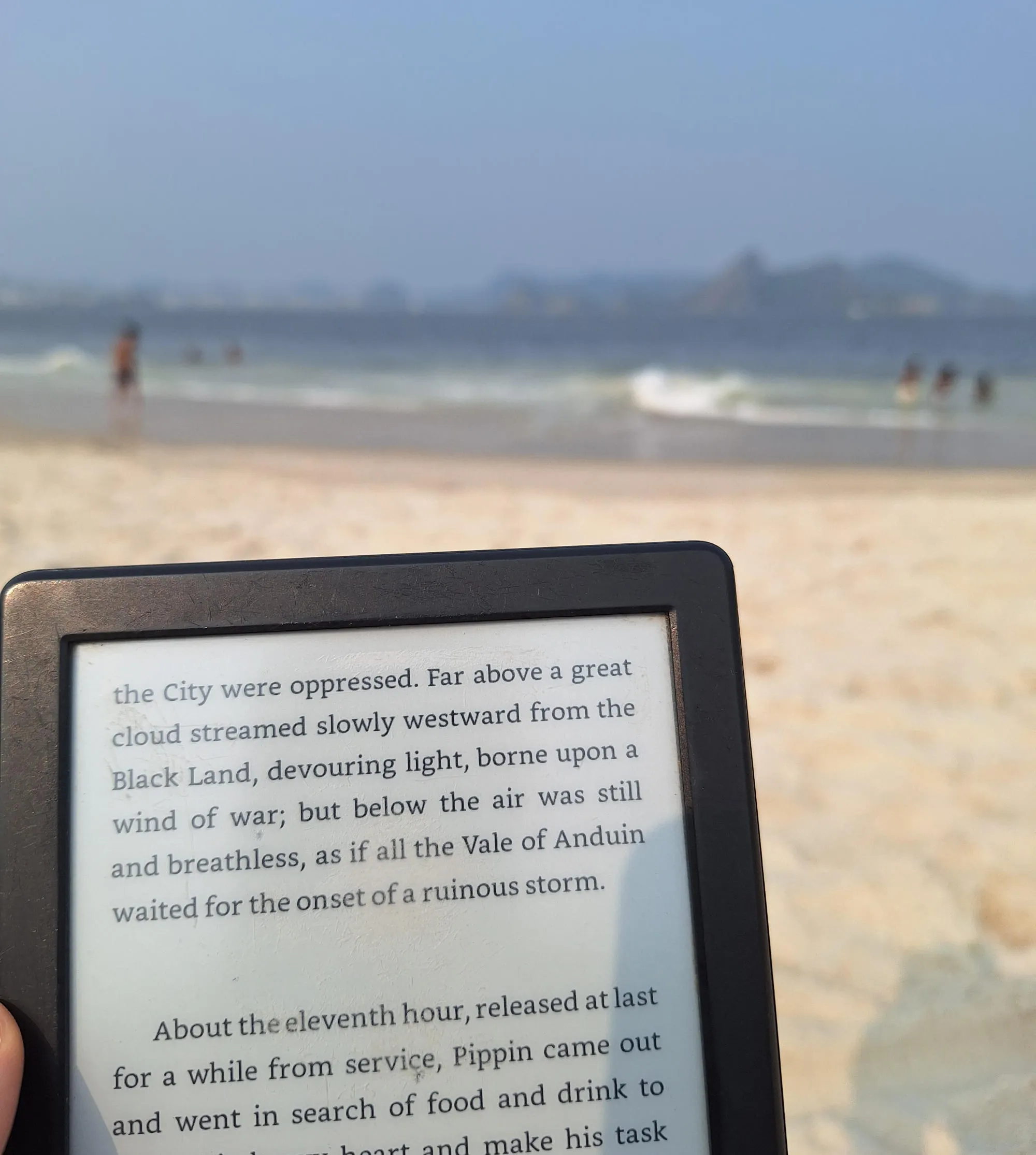 A close up photo of a Kindle e-book reader with a book open on it. The text is from Lord of the Rings. The background is out of focus, depicting a beach with the ocean and blue sky in the background.