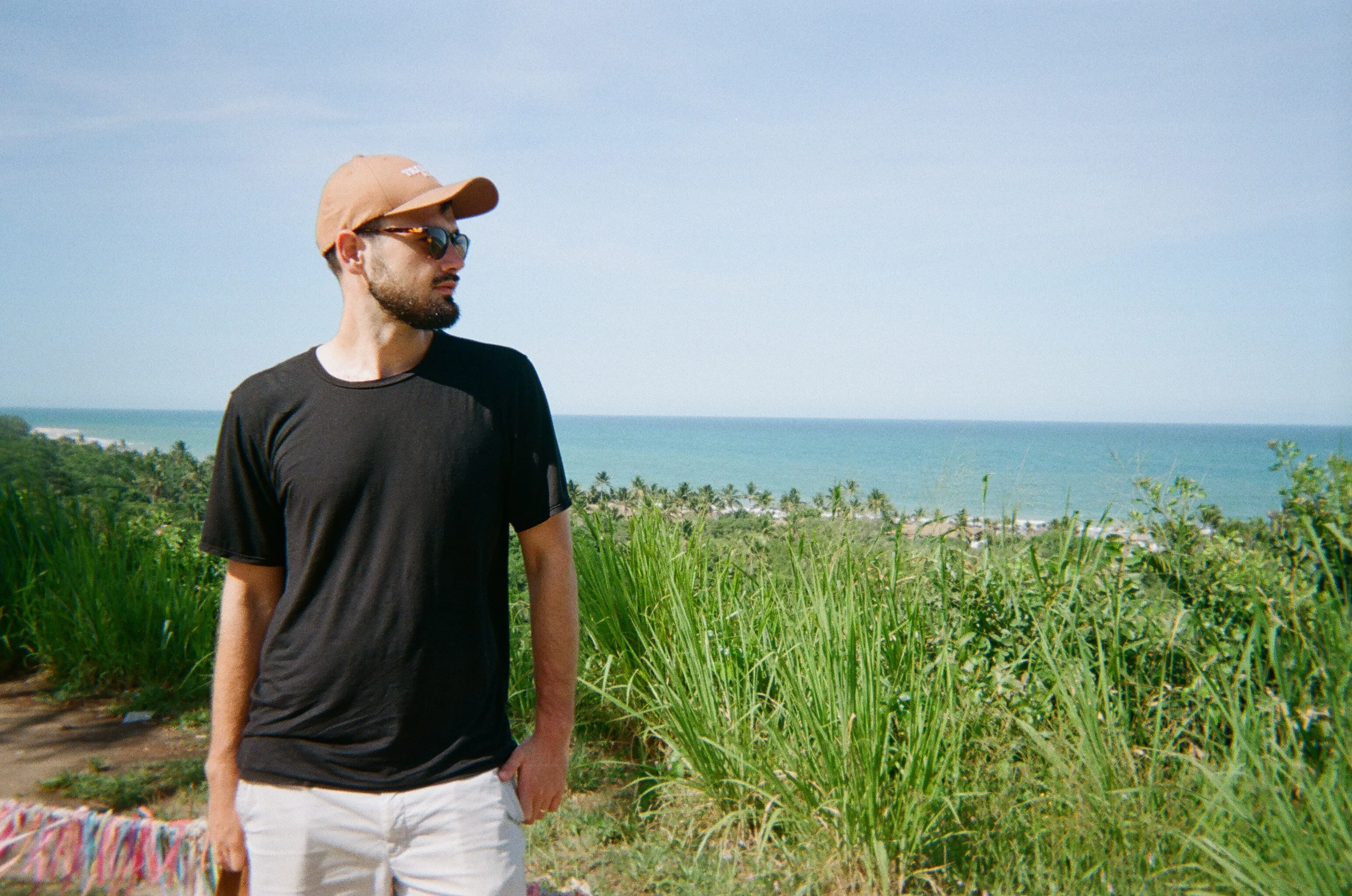 A film photo of me in sunglasses, wearing a black t-shirt and tan cap, standing in front of lush greenery with the ocean and blue sky in the background.