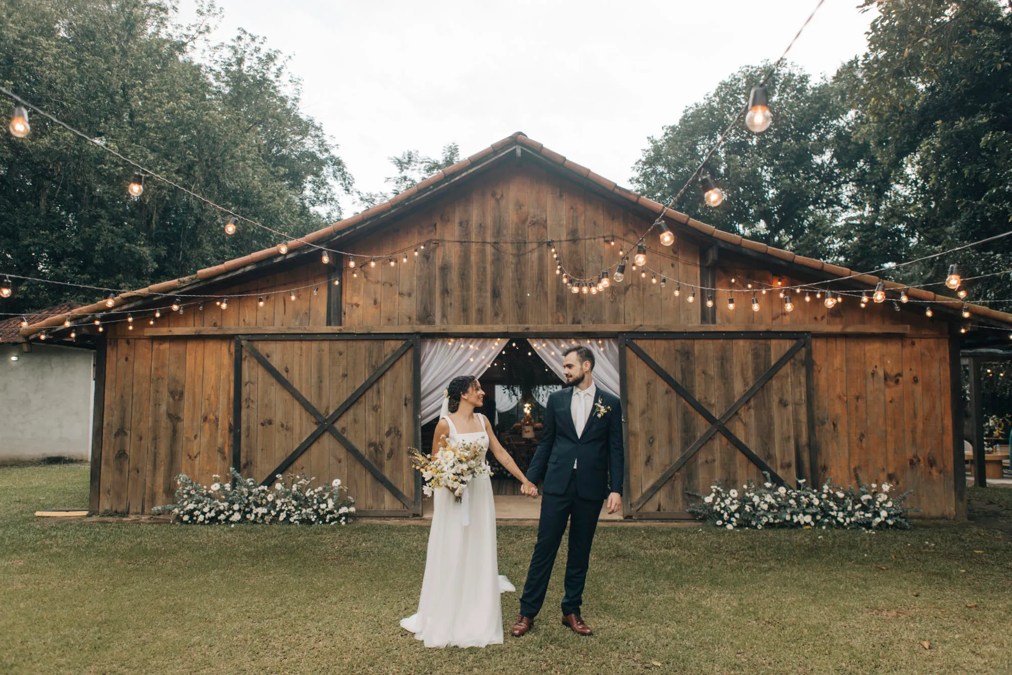 Me and my newlywed wife holding hands in front of a rustic wooden barn decorated with string lights and flowers, surrounded by greenery.